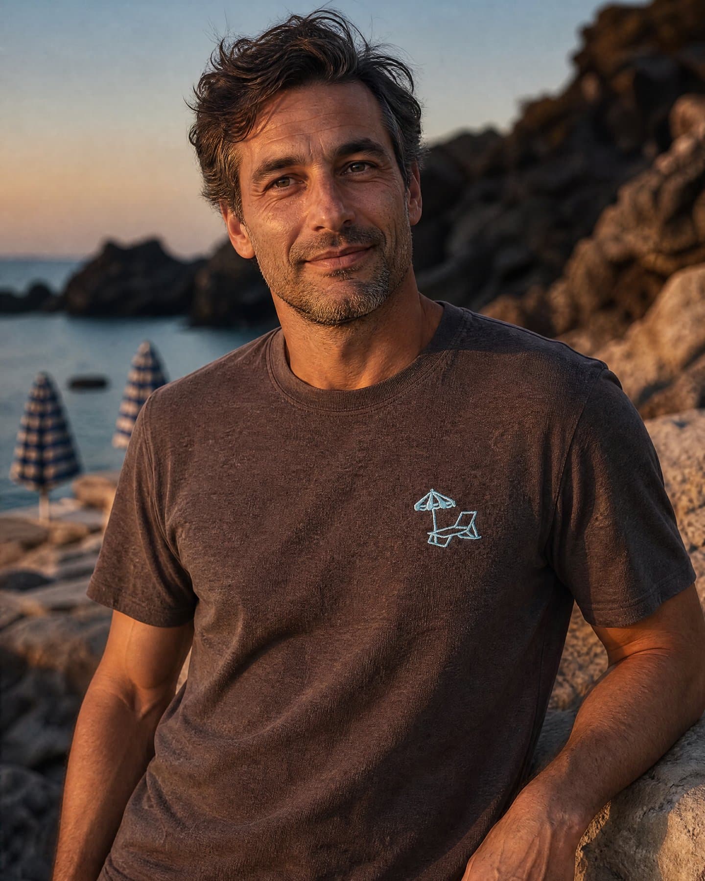Ischia Embroidery Tee worn on the rocks at golden hour, light blue chest embroidery visible