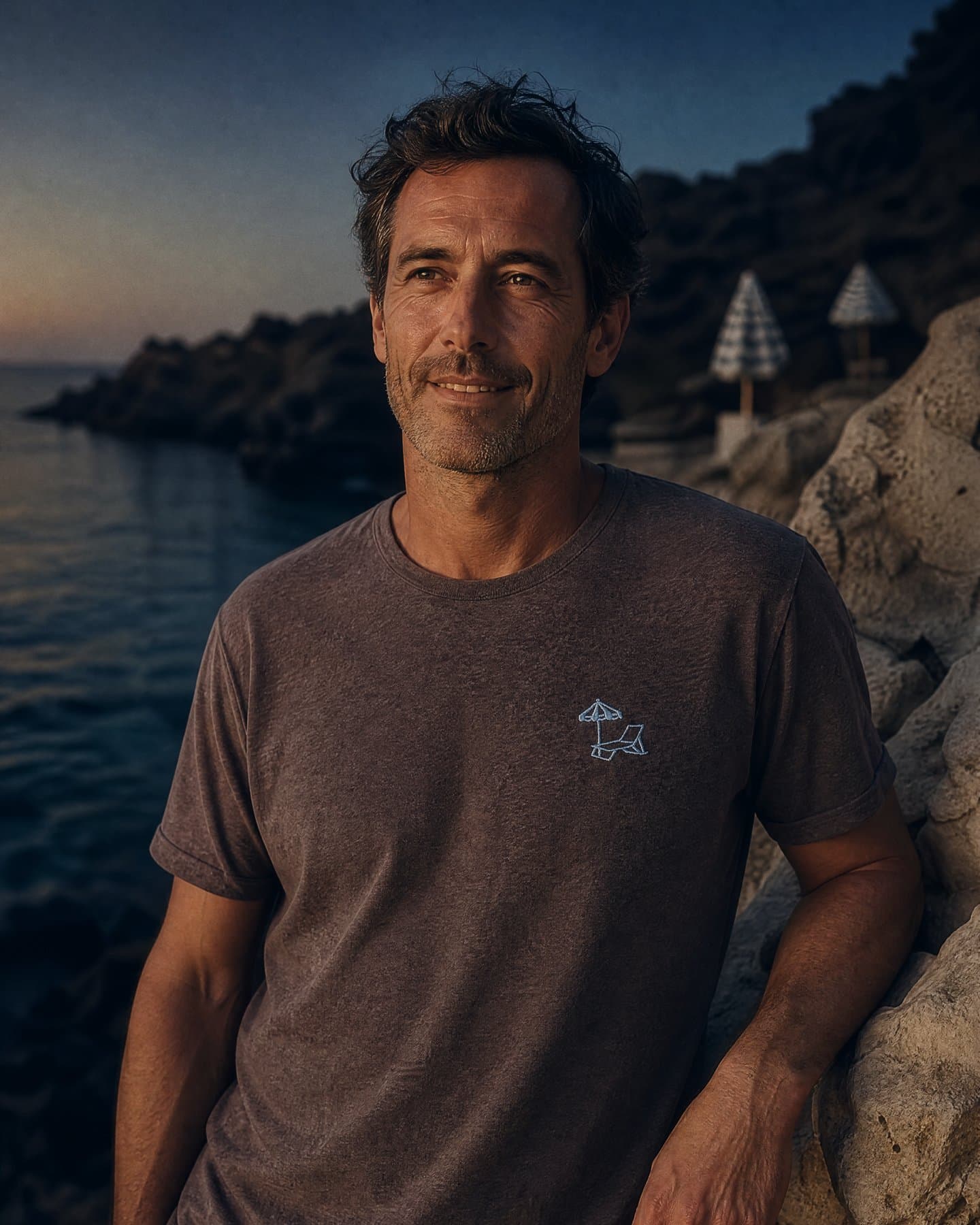 Ischia Embroidery Tee worn against the rocks at dusk, light blue chest embroidery visible